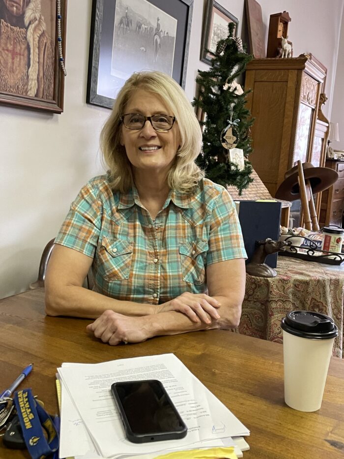 An older woman sits at a table smiling. She wears glasses and a checker patterned shirt that is blue and tan.