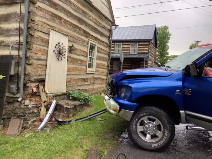 A blue truck with a dent in it sits outside a wooden house with a corner of its foundation destroyed. It is the aftermath of a collision with the building.