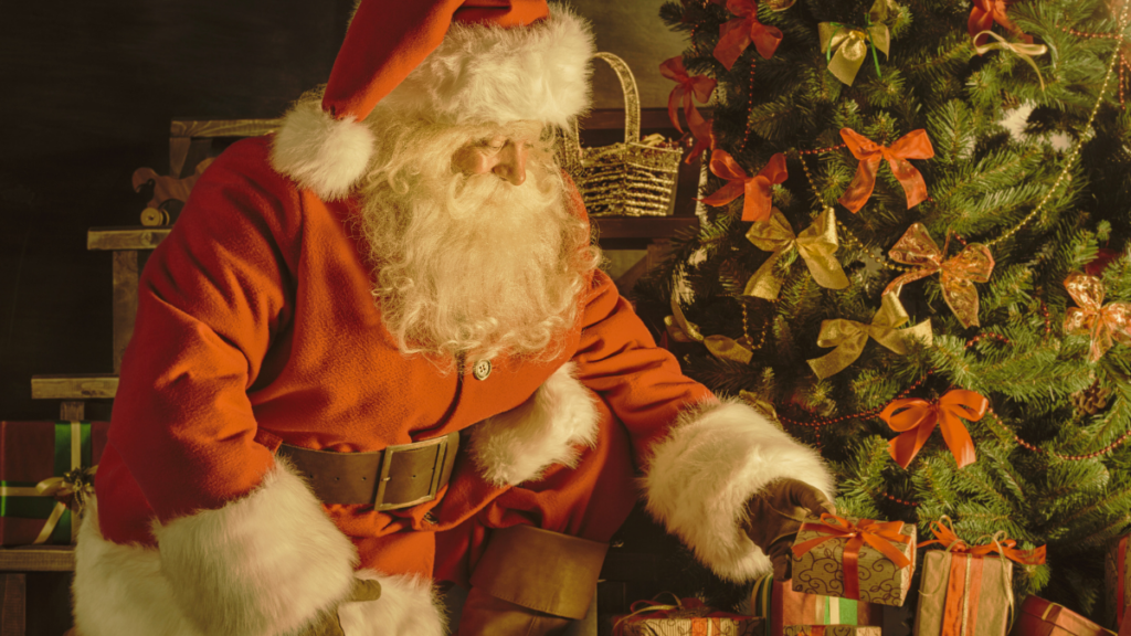 A man with a white beard and red suit bends down toward the bottom of a Christmas tree. He places a gift underneath its branches.