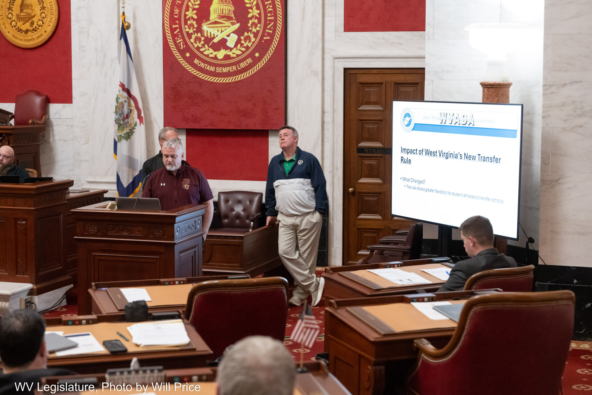 A man in a maroon sweater speaks at a lectern in a grand room paneled in white marble. Directly behind him stand two other men and arrayed in front of him are several wooden desks, a few occupied by state senators.