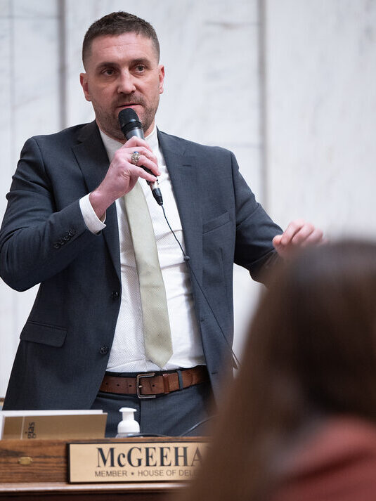 A man in a suit and tie standing behind a wooden desk speaks into a microphone and gestures with his left hand.