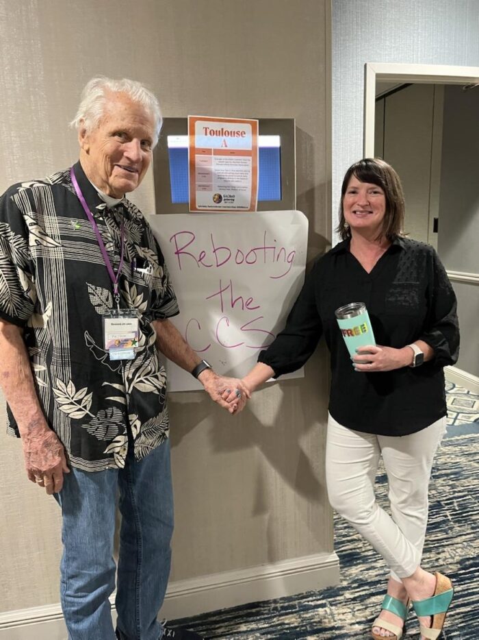 An elderly man holds hands with a woman with brown hair. Behind them is a sign that reads, "Rebooting the CCS." The woman is also holding a travel mug that says, "FREE" on it.