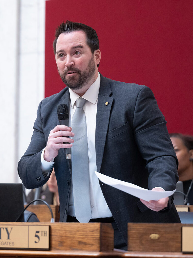 A man in a suit and tie stands behind a wooden desk with a name placard that reads "Fluharty, Member, House of Delegates." He is holding a microphone in his right hand and speaking into it. In his left hand, he is holding a sheet of paper and gesturing.