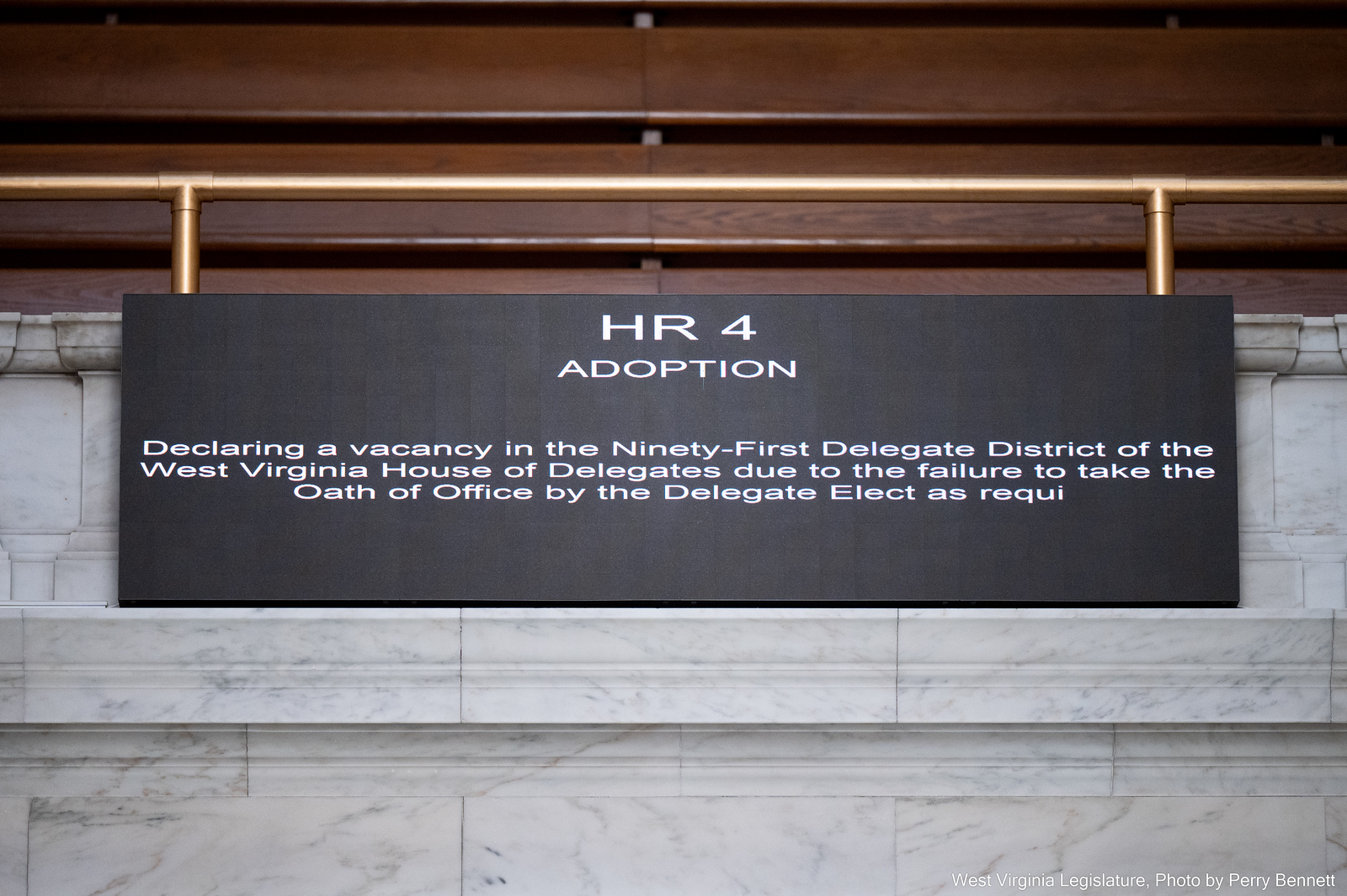 A black screen reads "HR 4 Adoption. Declaring a vacancy in the Ninety-First Delegate District of the West Virginia House of Delegates due to the failure to take the Oath of Office by the Delegate Elect as requi... ." The phrase is cut off. The screen is mounted on a marble wall.