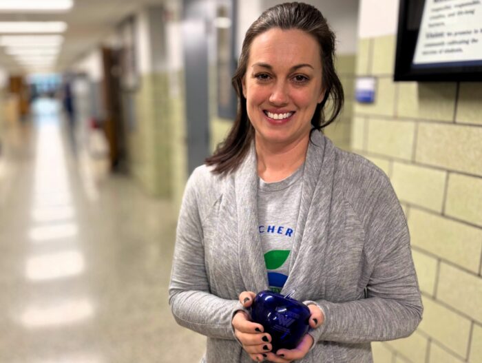 A woman with dark hair, wearing a gray sweater over a t-shirt, holds a blue apple Blenko glass paperweight. The woman stands in a school hallway.
