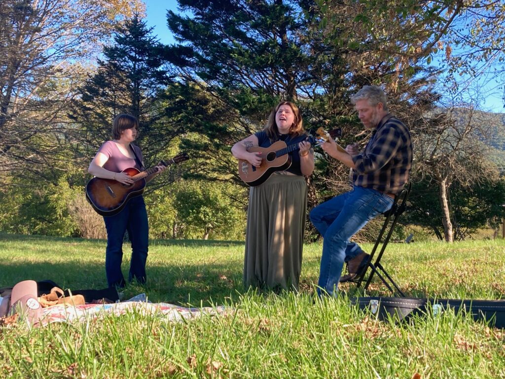 Three adults play musical instruments outside on a sunny day.