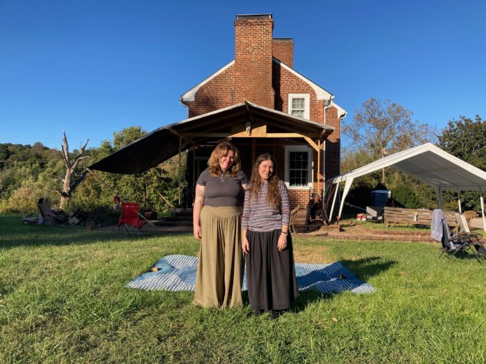 Two women stand outside on a sunny day in front of a brick house.