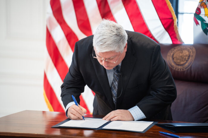 A man with white hair, glasses and wearing a dark suit stands at a desk in front of a large U.S. flag. He leans over the desk to sign papers in a folder using a blue pen.
