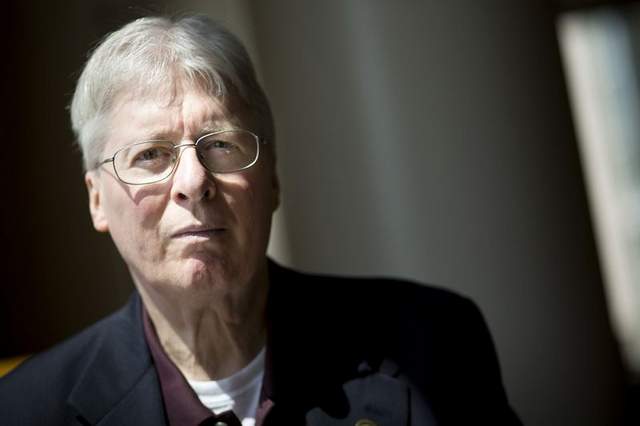 An older man with salt and pepper hair. He wears glasses with a navy blazer over a maroon shirt. Underneath the maroon shirt is a white shirt.