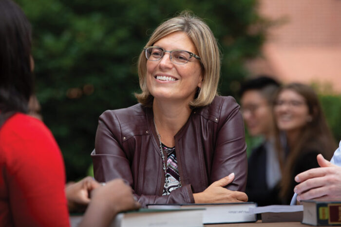 A woman with short, dark blond hair at the shoulder. She sits at a table and appears to be talking with others there who are unidentifiable. She wears a leather jacket, glasses, and a shirt featuring a black and white design.