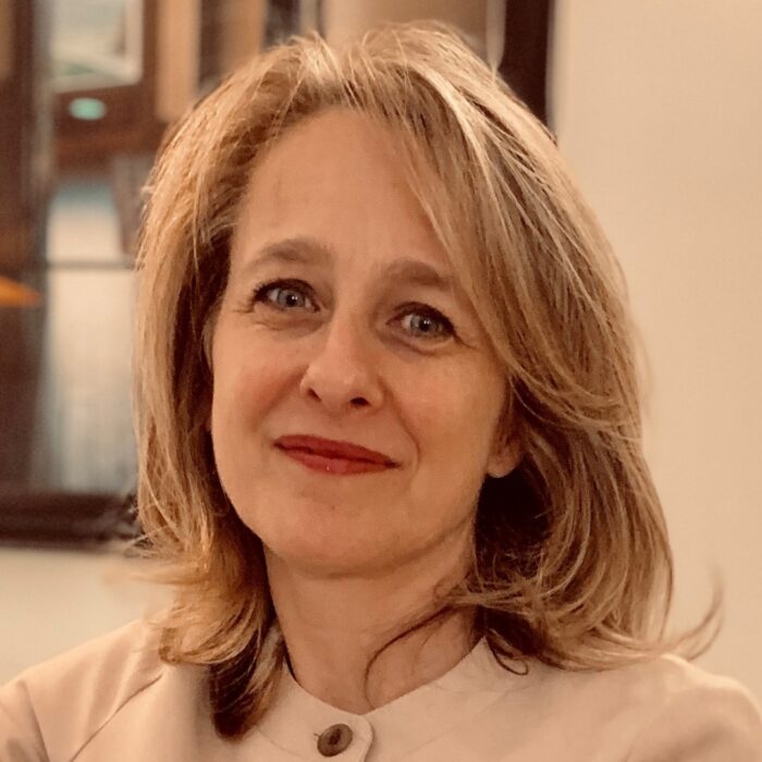 A headshot of a woman. He has blond hair and smiles for the camera. She wears an off-white button up shirt or jacket.