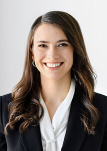 A headshot of a woman with long dark hair. She smiles for the camera and wears a white button up shirt with a black jacket over top. She is wearing small hoop earrings.