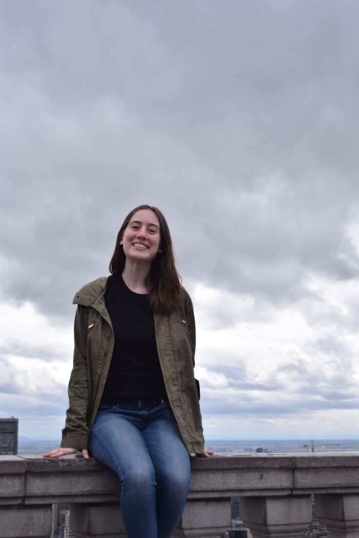 A woman sits on the stone siding of a balcony or bridge. She smiles as an overcast sky is seen above her. She wears blue jeans, a black shirt, and a tan jacket. She has long dark hair.