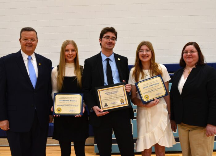 Five people stand in a line facing the camera for a photograph. The man on the left wears glasses and a light blue tie over a white button down shirt and a dark suit. To his right a young woman wears her blond hair down and a black apron dress over a white turtleneck while holding a certificate. To her right a young man wearing glasses and a black tie over a light blue button down shirt and a black suit also holds a certificate. To his right another young woman wears her blond hair down over a white dress, and she also holds a certificate towards camera. A woman stands at the right end of the line wearing a dark blue blazer over a white blouse and khaki pants.