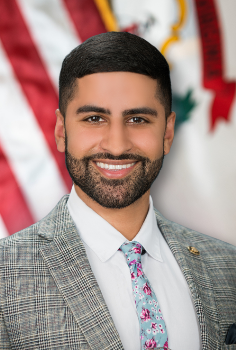 A man wearing a suit and tie poses in front of the U.S. and W.Va. flags. He has dark hair.