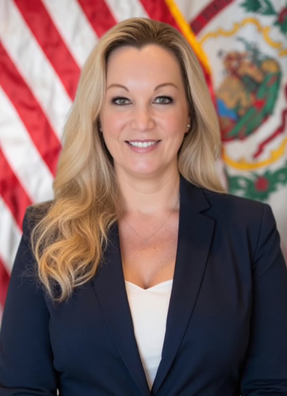 A woman wearing a suit poses in front of the U.S. and W.Va. flags. She has blonde hair.