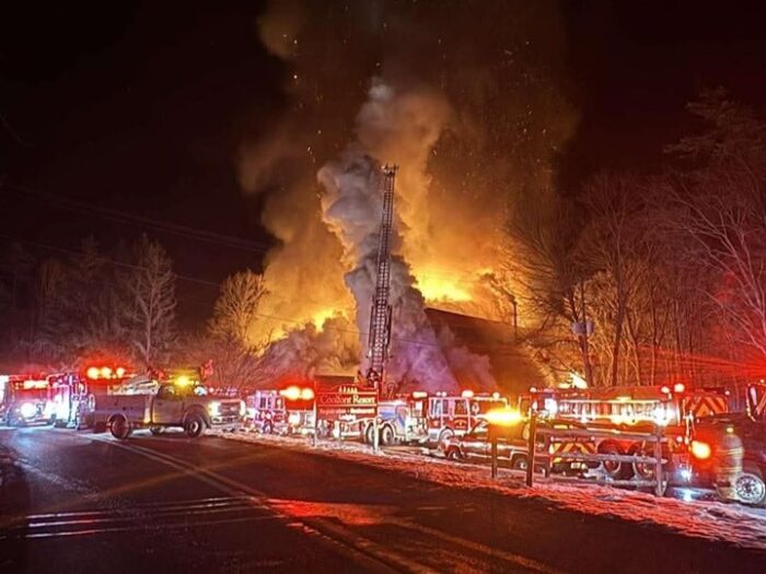 Dire trucks line the road outside a building engulfed in smoke and flames. Snow lies outside the building.