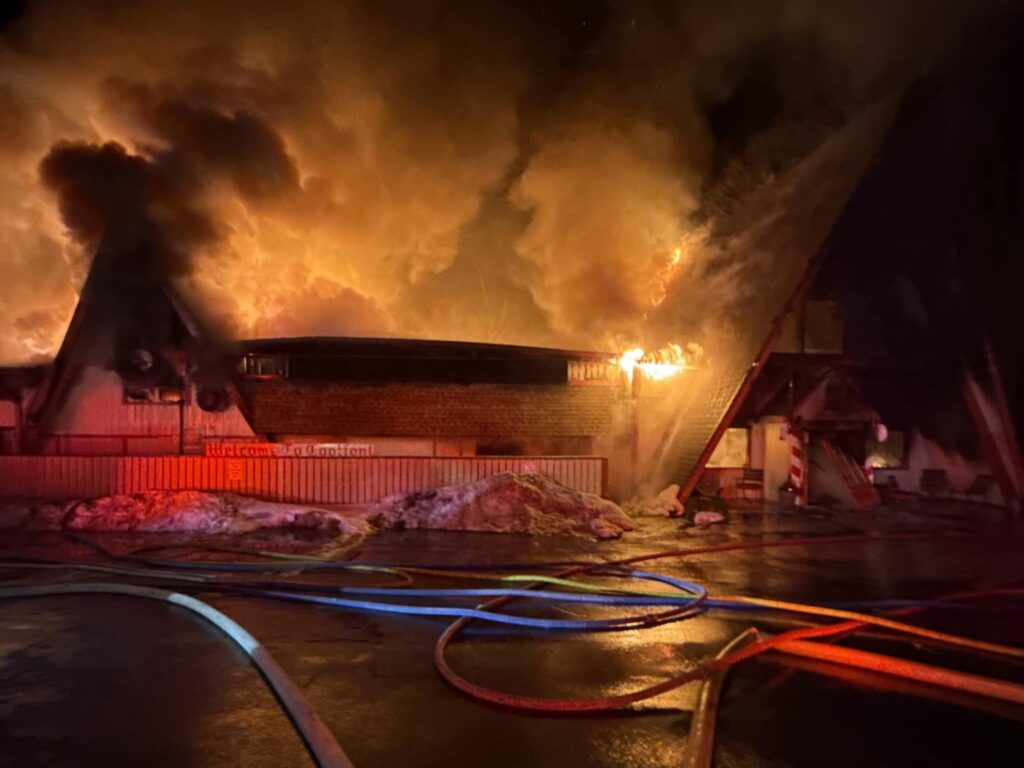 A flat building with triangular roofs is ablaze under the night sky. Snow sits out front of the structure, and numerous hoses stretch across the pavement toward the building.