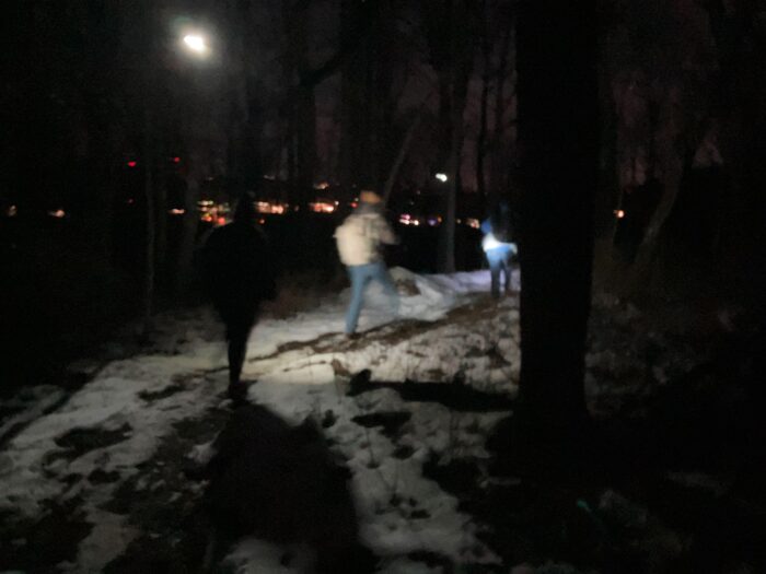 A light shines down on a snowy path in a wooded area at night. Three figures can be seen on the trail holding flashlights. Through the trees and in the far background can be seen orange lights.