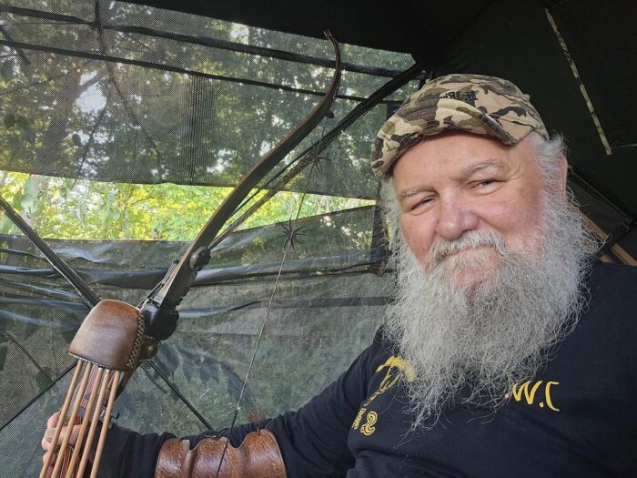 A man with a beard and camouflage hat holds a crossbow in the woods.