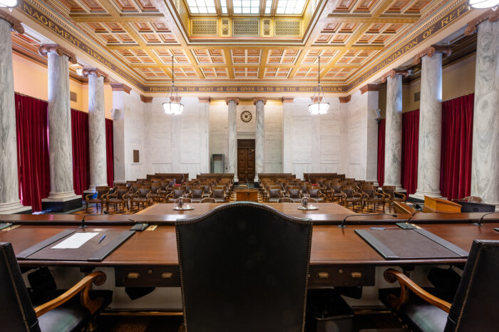 Rows of chairs sit empty before a court desk in a white marble courtroom with red velvet curtains.
