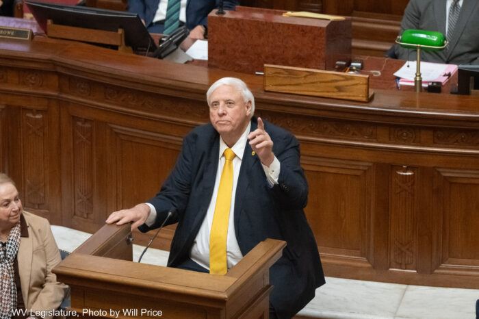 man in a large yellow tie points to the ceiling while speaking from behind a wood podium
