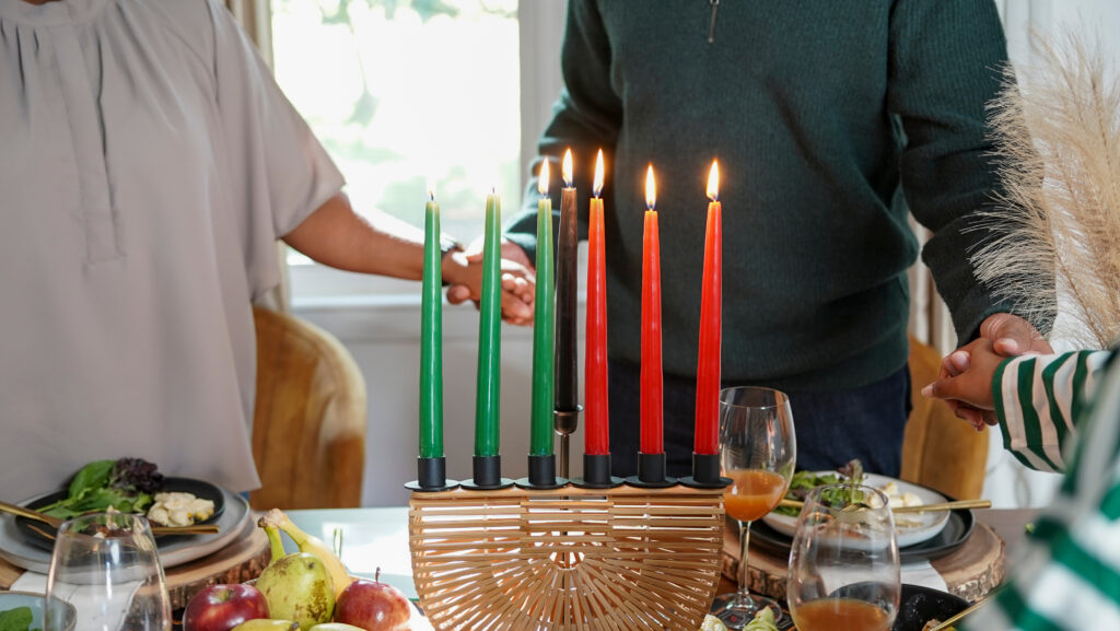 Family with daughter (12-13) holding hands over Kwanzaa meal. In center of frame is a wicker candelabra with three green candles, one black candle in the middle, and three red candles on the left.