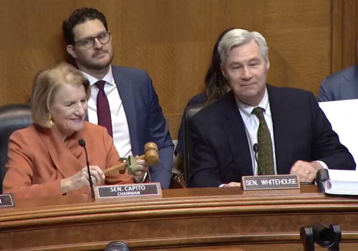 A woman in an orange jacket holds a wooden gavel next to a man in a dark suit and tie in a wood-paneled room with others present.