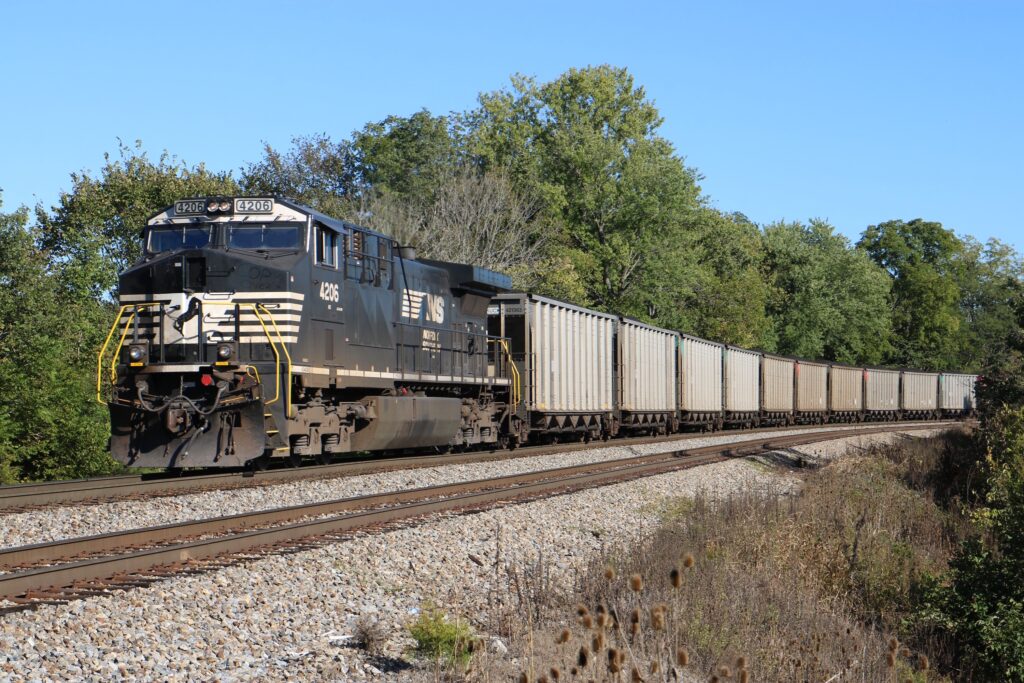 A black locomotive and a line of silver rail cars against the backdrop of late-summer foliage and a clear sky.