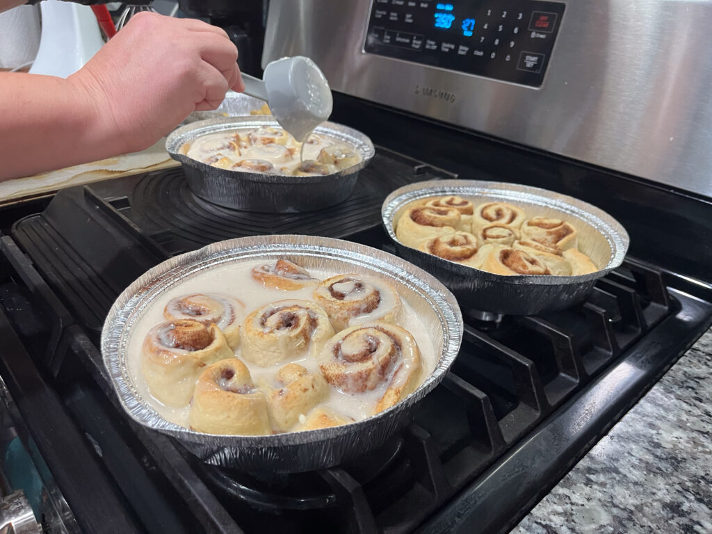 Three pans of cinnamon rolls are being prepared on the surface of a gas-top stove.