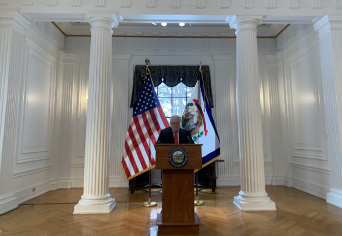 man stands at a podium in front of the American and West Virginia flags, speaking