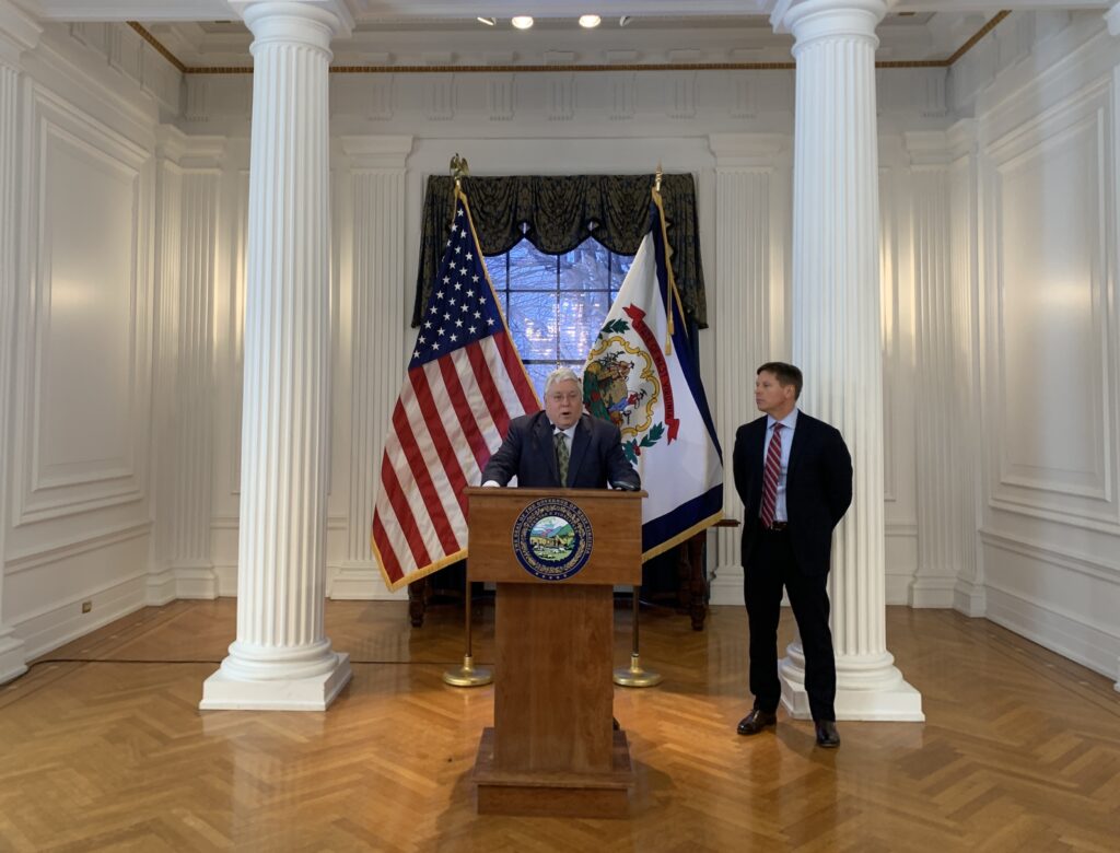 man speaks at a podium, another man stands to his left