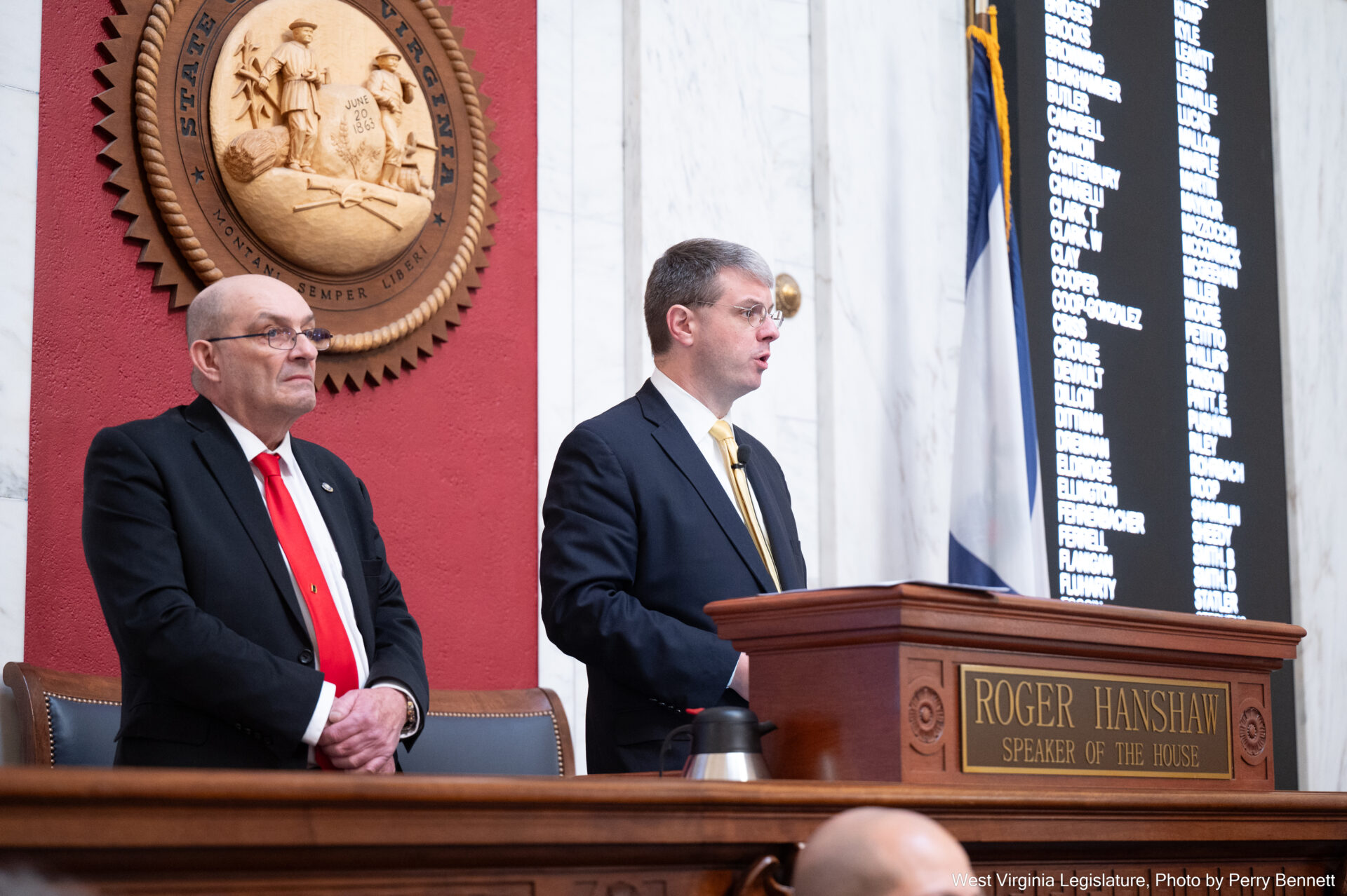 man with a yellow tie speaks at a podium, man with a red tie stands behind him