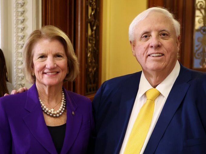 A woman in a purple suit with necklace and a man in navy suit with pale yellow tie smile at the camera.