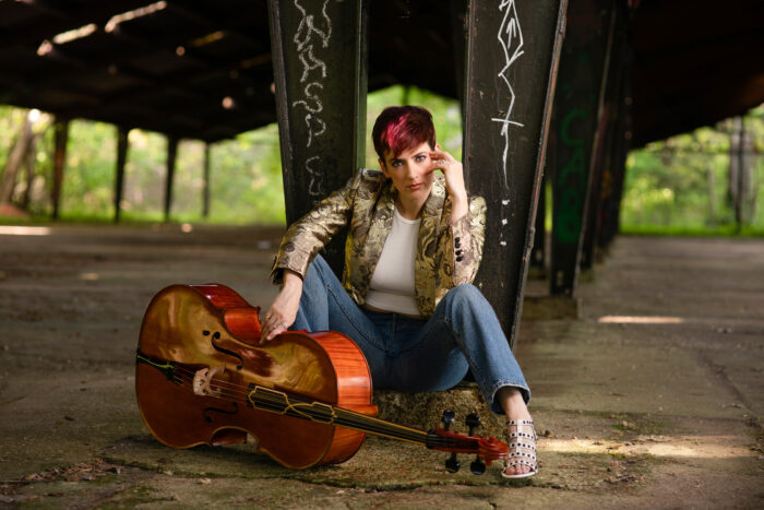 A woman is sitting on concrete under an old pavilion with a cello by her feet. She has short deep red hair with a pink streak in it. She is wearing a gold brocade jacket, a white shirt, jeans, and white studded sandal heels. Her hand is on her face.