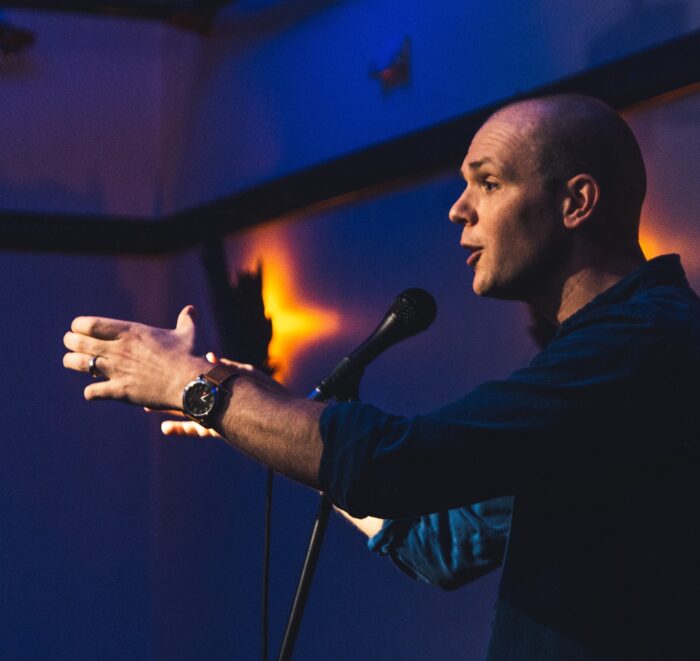 A man in a blue shirt talks into a microphone and gestures as he tells a story.