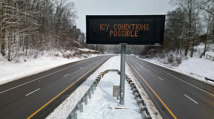 A two-way road has two lanes of traffic in each direction, separated by a median. An electric sign in the median reads "Icy Conditions Possible."