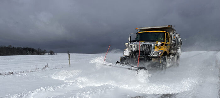 A snow plow pushes several inches of snow off of a rural roadway.