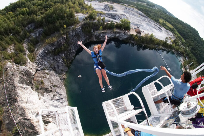 A woman with red hair and a blue shirt is freefalling toward a body of water in a quarry, attached to a platform above by a long blue elastic cord. A staff members stands on the platform above.