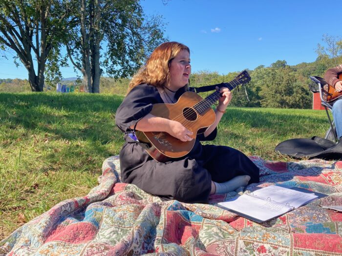 A woman with a guitar sits on a quilt near two trees and sings.