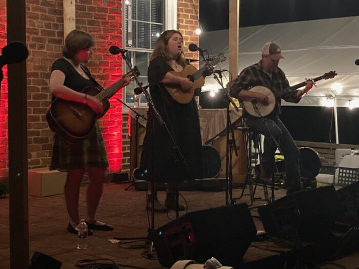 A trio of folk musicians stand on a porch and perform.