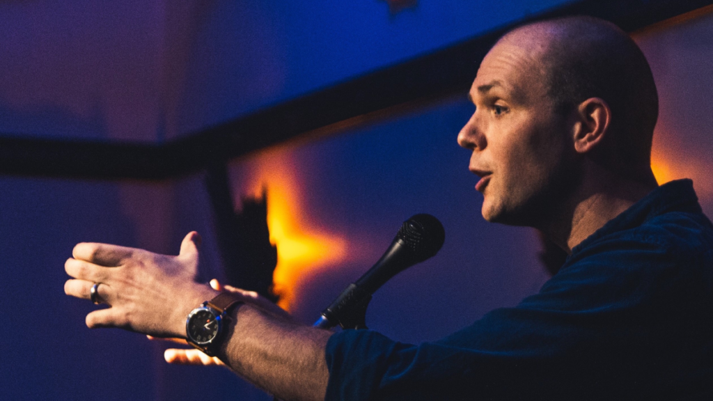 A man speaks into a microphone. He has his arms stretched out and is talking to an audience.