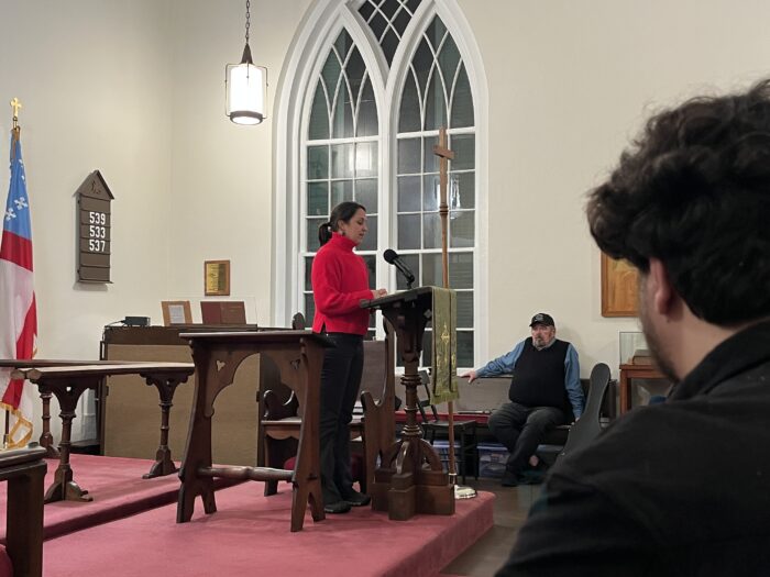 A woman in a red turtle stands on the podium in a church and talks in a microphone. The members of the audience look at her listening.