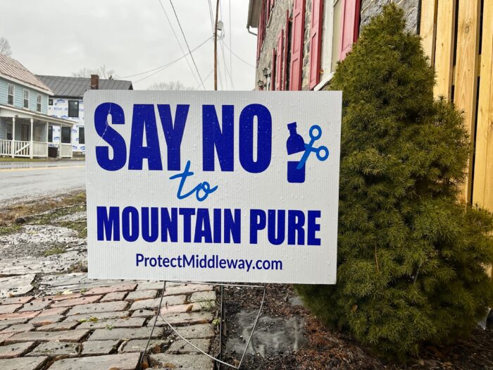 A sign on the street on the city street reads "Say no to Mountain Pure" In Blue Block text. He sits next to a stone path and a small bush.