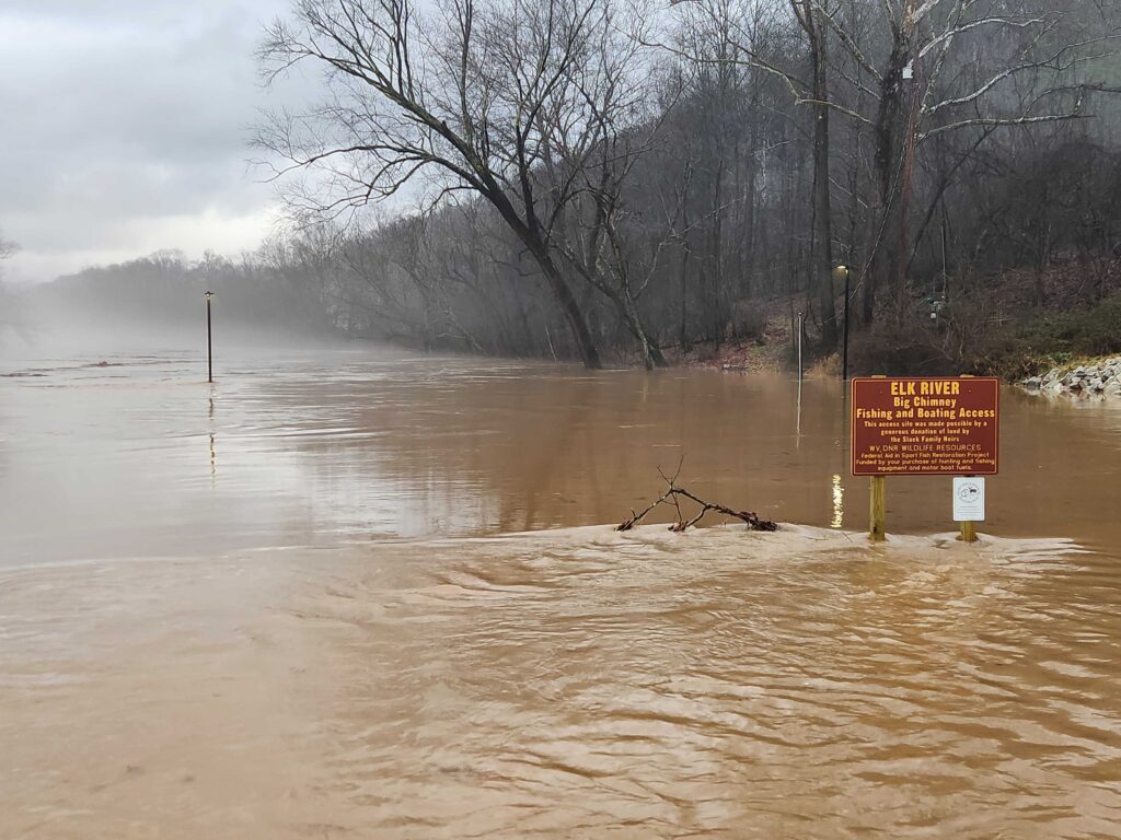 A brown sign sits above a flooded waterway and reads "Elk River." It is hard to delineate where the river lies, because flooding has raised water levels above the riverbank.