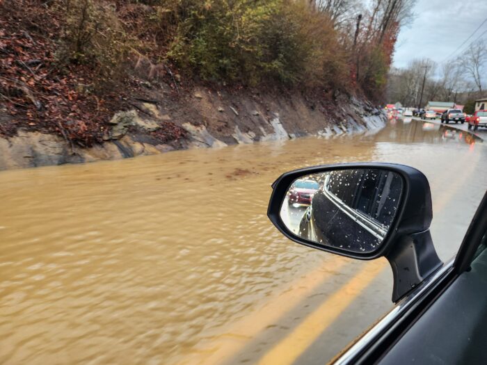 A two-lane asphalt road divided by double yellow lines shows standing water filling one lane near the slope of a hill. Cars wait in line in the other lane.