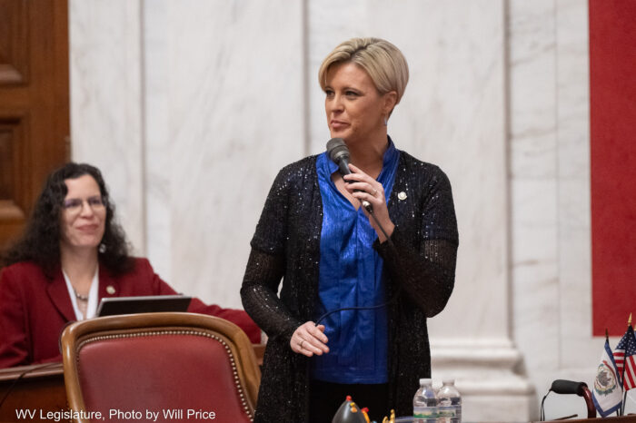 A woman weaking a sparkling knee length sheer black sweater over a satin royal blue blouse holds a microphone while standing in front of a chair with red leather lining. Behind her can be seen a woman seated at a wooden desk wearing a burgundy blazer over a white blouse. In the background a white marble wall has a column facade.