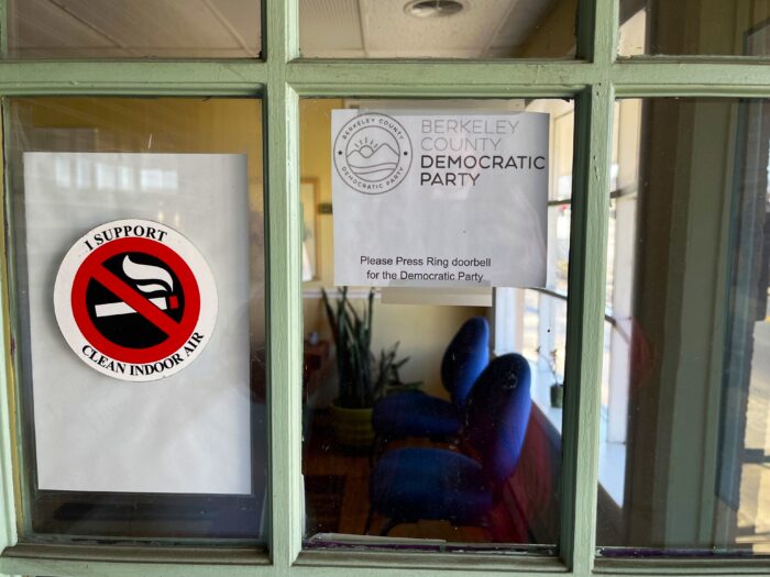 Viewed from close up, glass window panes on a door are split by green-painted wood. A piece of paper taped to the window reads "Berkeley County Democratic Party." A waiting room and house plant are visible in the building's interior through the window.