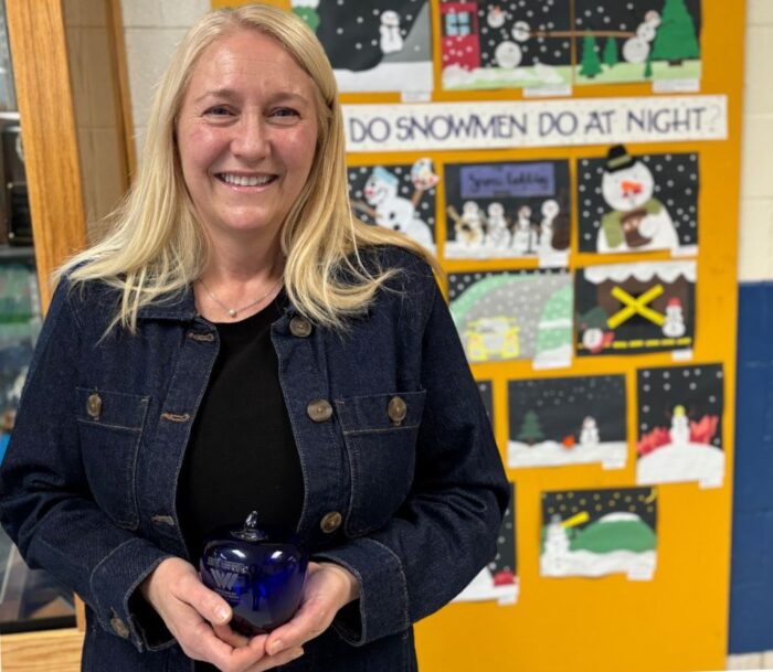 A woman with long blond hair wearing a jean jacket poses in front of a poster. She holds a blue, glass apple paperweight made by Blenko Glass Company. She is smiling.