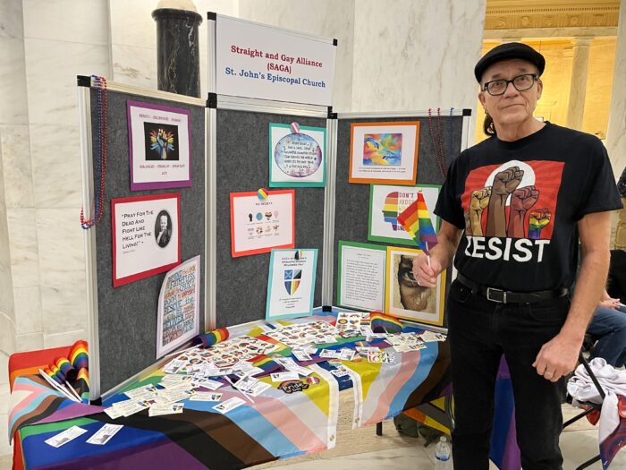 A man holds a rainbow LGBTQ Pride flag and stands in front of a table lined with stickers and Pride flag tablecloths. On top of the table is a display that reads "Straight and Gay Alliance." The man is wearing a shirt with several illustrated fists raised in the air and the text "RESIST." The setup is located in a white marble hallway.